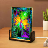 A stained-glass-style decorative light box displaying a colorful fairy design, standing on a wooden surface next to a small potted plant.