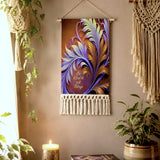 A  fringed canvas decorative wall hanging with a colorful abstract feather design in purple, blue, and gold, hanging on a beige wall in a styled room with plants and a macrame wall hanging. Inspirational text Be Kind to All Things