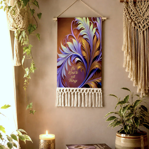 A  fringed canvas decorative wall hanging with a colorful abstract feather design in purple, blue, and gold, hanging on a beige wall in a styled room with plants and a macrame wall hanging. Inspirational text Be Kind to All Things