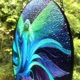 Side view showing texture of a Round glass fairy suncatcher in a black frame and a hanging black chain featuring Lunaria Silvermist of the Fae Realm by Primal Painter. Blues, greens, and purples, and florals. 