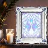 Soft violet and white angel with blue orbs in her hands and above her head surrounded by symmetrical energy patterns. Art called On the Wings of Angels by Primal Painter. Framed on a shelf with candles
