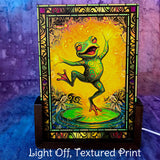 Small acrylic table lamp with the light off featuring a dancing frog artwork  in oranges, greens, and reds with a stained glass-effect border. Art by Primal Painter