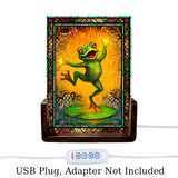 Small acrylic table lamp featuring a dancing frog artwork  in oranges, greens, and reds with a stained glass-effect border. USB plug, adapter not included text.  Art by Primal Painter