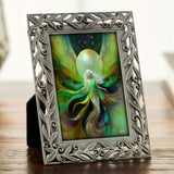 An ornate silver picture frame with a leafy scroll border, holding a fantasy art print of a glowing green  and gold fairy with a large moon, displayed on a wooden surface.  Art by Primal Painter