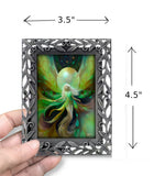 A hand holds a small decorative picture frame against a white background. The frame is ornate and metallic gray, containing a fantasy artwork of a winged fairy with green and gold tones.