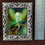 A framed fantasy art print featuring a glowing fairy with green and yellow wings, shown next to a metal ruler for scale on a brown textured background.