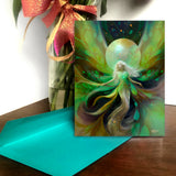 A story card featuring a fantasy illustration of a glowing fairy goddess with flowing green and gold hair, placed on a wooden surface next to a bright turquoise envelope. A vase with green stems and a pink ribbon is blurred in the background.