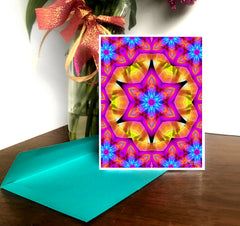 A colorful mandala art greeting card with a star and geometric pattern, displayed on a desk with a decorative ribbon and a teal envelope by Primal Painter.