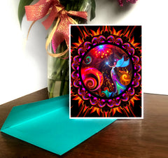 A greeting card with a colorful mandala design and an angel motif, with a red and orange color scheme, displayed on a table with a blue envelope. Art by Primal Painter