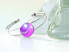 A silver cuff bracelet with a violet-colored swirl design, displayed on a glass surface by Primal Painter.